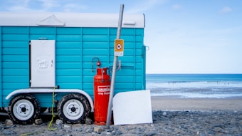 A turquoise trailer with white doors and wheels is parked on a rocky beach. A red propane gas cylinder and a metal pole with a sign are placed in front of the trailer. In the background, the ocean waves gently roll onto the shore under a partly cloudy sky.