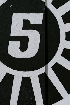 A bold, stylized black and white mural featuring a large number five in the center. Radiating lines extend outward from the circle encompassing the number, creating a dynamic, sunburst-like design.