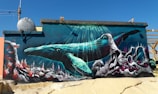 A vibrant mural depicts an underwater scene featuring a large blue whale swimming among colorful coral reefs. Sunlight pierces the water, illuminating the whale and the intricate details of the underwater environment. Various signs with messages such as 'Be The Change' and 'Wave of Change' are integrated into the artwork.
