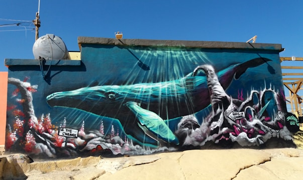 A vibrant mural depicts an underwater scene featuring a large blue whale swimming among colorful coral reefs. Sunlight pierces the water, illuminating the whale and the intricate details of the underwater environment. Various signs with messages such as 'Be The Change' and 'Wave of Change' are integrated into the artwork.