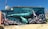 A vibrant mural depicts an underwater scene featuring a large blue whale swimming among colorful coral reefs. Sunlight pierces the water, illuminating the whale and the intricate details of the underwater environment. Various signs with messages such as 'Be The Change' and 'Wave of Change' are integrated into the artwork.