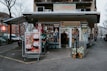 A small grocery store is located on a city street corner, with a mix of international and local products visible through the entrance. The storefront is decorated with colorful posters featuring fruits and vegetables, and mannequins dressed in traditional outfits. Outside, there are two carts filled with fresh produce like cabbages and packaged goods. A person is standing in the entrance, possibly a customer or a store attendant. Nearby buildings and parked cars are part of an urban neighborhood setting.