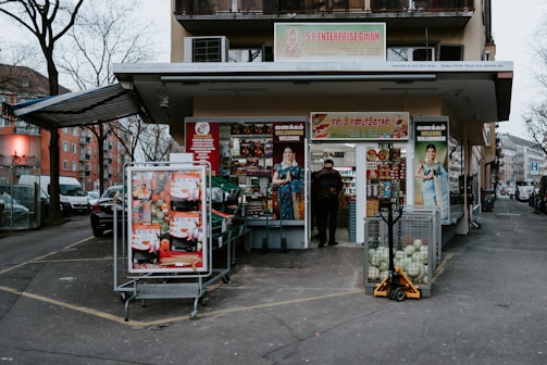 A small grocery store is located on a city street corner, with a mix of international and local products visible through the entrance. The storefront is decorated with colorful posters featuring fruits and vegetables, and mannequins dressed in traditional outfits. Outside, there are two carts filled with fresh produce like cabbages and packaged goods. A person is standing in the entrance, possibly a customer or a store attendant. Nearby buildings and parked cars are part of an urban neighborhood setting.