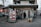 A small grocery store is located on a city street corner, with a mix of international and local products visible through the entrance. The storefront is decorated with colorful posters featuring fruits and vegetables, and mannequins dressed in traditional outfits. Outside, there are two carts filled with fresh produce like cabbages and packaged goods. A person is standing in the entrance, possibly a customer or a store attendant. Nearby buildings and parked cars are part of an urban neighborhood setting.