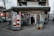 A small grocery store is located on a city street corner, with a mix of international and local products visible through the entrance. The storefront is decorated with colorful posters featuring fruits and vegetables, and mannequins dressed in traditional outfits. Outside, there are two carts filled with fresh produce like cabbages and packaged goods. A person is standing in the entrance, possibly a customer or a store attendant. Nearby buildings and parked cars are part of an urban neighborhood setting.