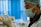 A healthcare professional wearing a surgical mask, disposable gloves, and a hairnet closely examines a vial of liquid. The background is blurred with blue and white tones, suggesting a clinical environment.