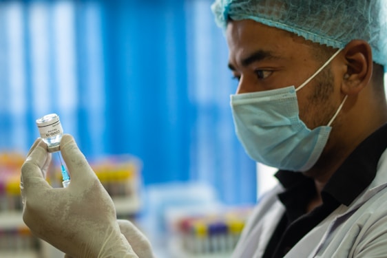 A healthcare professional wearing a surgical mask, disposable gloves, and a hairnet closely examines a vial of liquid. The background is blurred with blue and white tones, suggesting a clinical environment.