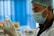 A healthcare professional wearing a surgical mask, disposable gloves, and a hairnet closely examines a vial of liquid. The background is blurred with blue and white tones, suggesting a clinical environment.