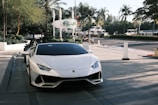 A sleek white sports car is parked on a paved driveway, positioned towards lush green palm trees in the background. A sign reading 'Eden Roc' appears behind the car, set against a backdrop of yachts and a calm street scene. The scene conveys luxury and leisure with modern urban elements.