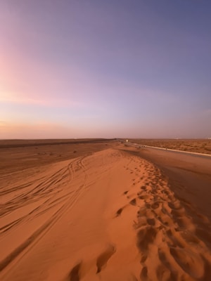 A vast desert landscape with orange sand dunes extending into the distance, featuring patterns of vehicle tracks and footprints. The sky above is expansive with soft gradients of blue and pink hues, suggesting a time of sunset or sunrise.