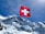 A Swiss flag waves prominently against a backdrop of majestic snow-covered mountains under a clear blue sky. The mountain peaks are rugged with visible glaciers.