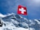 A Swiss flag waves prominently against a backdrop of majestic snow-covered mountains under a clear blue sky. The mountain peaks are rugged with visible glaciers.