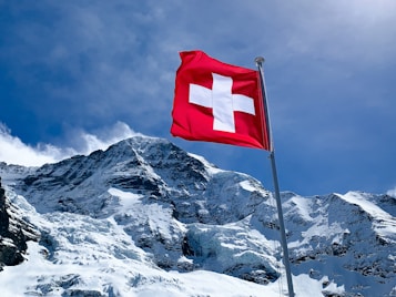 A Swiss flag waves prominently against a backdrop of majestic snow-covered mountains under a clear blue sky. The mountain peaks are rugged with visible glaciers.