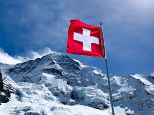 A Swiss flag waves prominently against a backdrop of majestic snow-covered mountains under a clear blue sky. The mountain peaks are rugged with visible glaciers.
