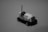 A black and white photograph of a small toy vehicle resembling a vintage tow truck. The toy is positioned on a plain surface with its front facing the viewer. The truck features detailed elements, including a cabin, grill, and a towing hook at the back.