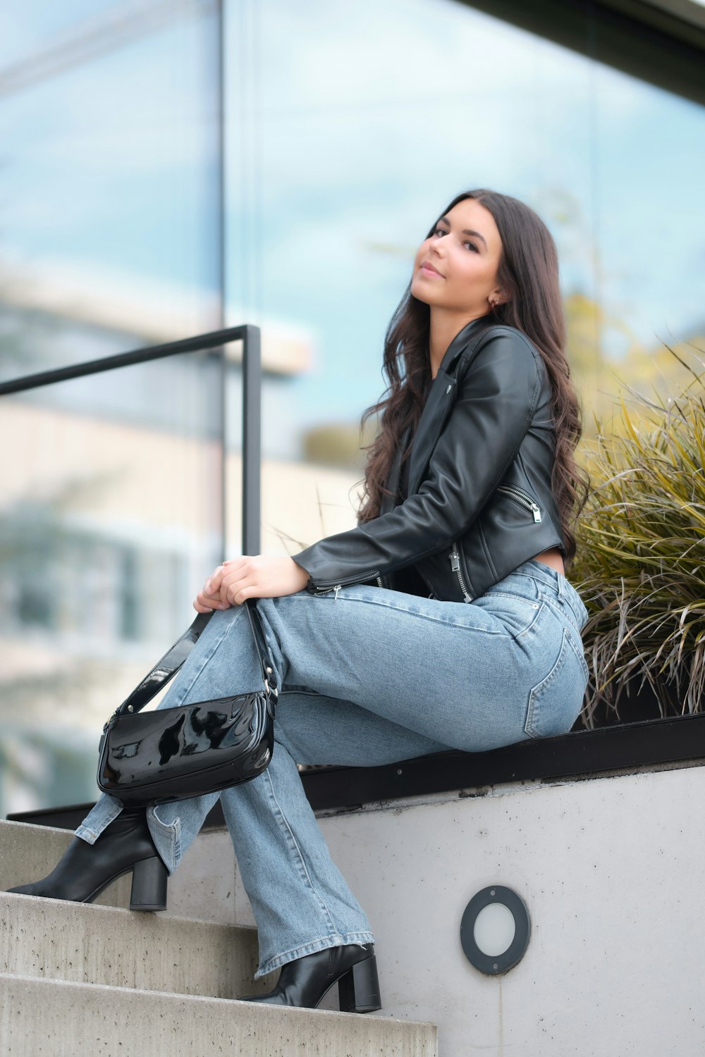 woman in black leather jacket and blue denim jeans sitting on white concrete bench during daytime