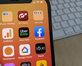 A smartphone displays a variety of app icons against a warm gradient background. The apps include Facebook, Uber Eats, Analytics, and others. In the background, a computer keyboard is visible on the desk.
