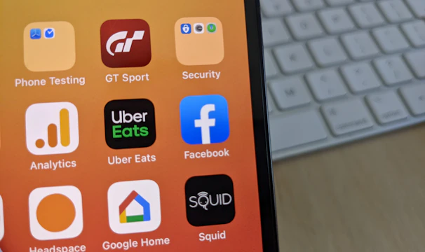 A smartphone displays a variety of app icons against a warm gradient background. The apps include Facebook, Uber Eats, Analytics, and others. In the background, a computer keyboard is visible on the desk.