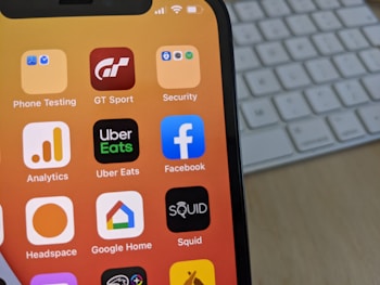 A smartphone displays a variety of app icons against a warm gradient background. The apps include Facebook, Uber Eats, Analytics, and others. In the background, a computer keyboard is visible on the desk.