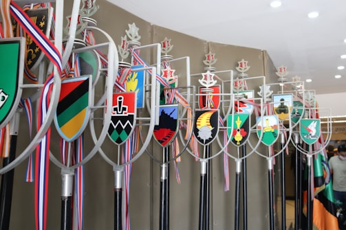 A collection of ceremonial staffs are adorned with various colorful heraldic shields. Each shield features unique symbols and designs, and they are topped with metal emblems. The staffs are arranged in a row, and ribbons in red, white, and blue are draped from them. The setting appears to be indoors with bright lighting overhead.