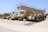 A row of military vehicles with crawler tracks are parked on a gravel surface. The vehicles are painted in desert camouflage colors, and one features a large, elevated structure resembling a bridge launcher. There are informational plaques in front of each vehicle.