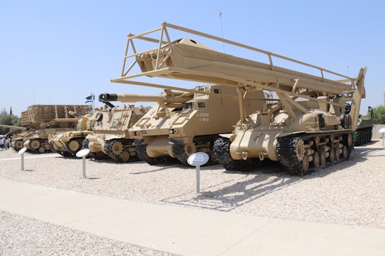 A row of military vehicles with crawler tracks are parked on a gravel surface. The vehicles are painted in desert camouflage colors, and one features a large, elevated structure resembling a bridge launcher. There are informational plaques in front of each vehicle.