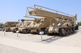 A row of military vehicles with crawler tracks are parked on a gravel surface. The vehicles are painted in desert camouflage colors, and one features a large, elevated structure resembling a bridge launcher. There are informational plaques in front of each vehicle.