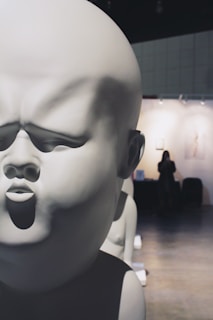A large, smooth, white sculpture of a baby's face, with exaggerated features and a closed-eye expression, fills most of the foreground. In the background, a small figure stands in an art gallery or exhibition space with dim lighting and framed artworks on the walls.