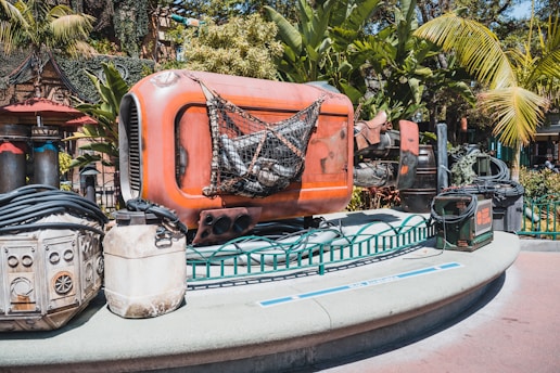 A futuristic-looking, orange metal contraption with industrial details is surrounded by lush, tropical plants. It sits on a platform with additional equipment, cables, and containers scattered around.