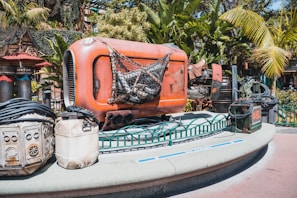 A futuristic-looking, orange metal contraption with industrial details is surrounded by lush, tropical plants. It sits on a platform with additional equipment, cables, and containers scattered around.