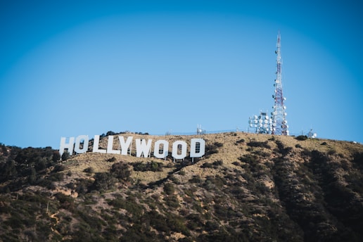 A large iconic sign spelling 'Hollywood' is situated on a hillside covered with sparse vegetation and dark green shrubs. Next to the sign, a tall communications tower with numerous satellite dishes is visible against a clear blue sky.