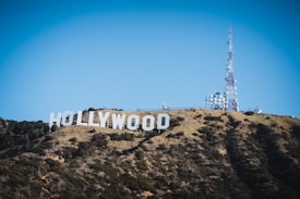 A large iconic sign spelling 'Hollywood' is situated on a hillside covered with sparse vegetation and dark green shrubs. Next to the sign, a tall communications tower with numerous satellite dishes is visible against a clear blue sky.