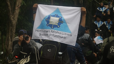 A person is holding a flag with a logo and the text 'Himpunan Mahasiswa Sistem Informasi'. People in graduation gowns and medals are gathered, with one individual photographing the scene. The environment is outdoors with trees visible in the background.