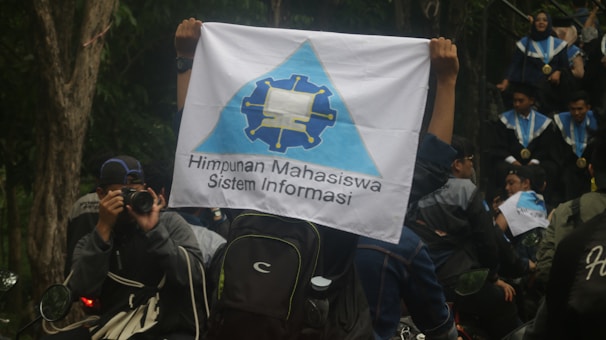 A person is holding a flag with a logo and the text 'Himpunan Mahasiswa Sistem Informasi'. People in graduation gowns and medals are gathered, with one individual photographing the scene. The environment is outdoors with trees visible in the background.