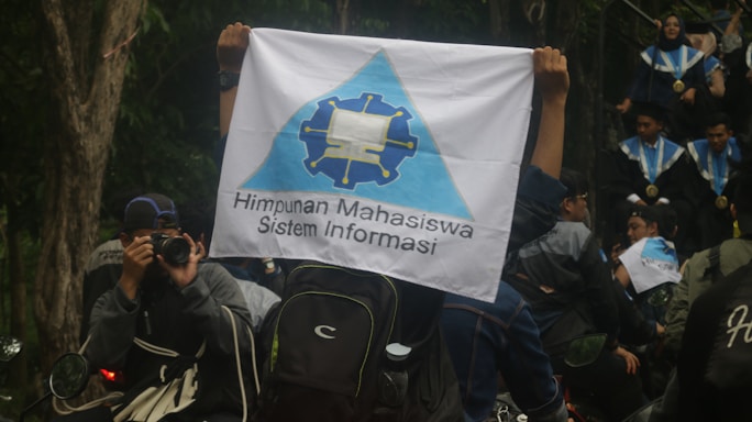 A person is holding a flag with a logo and the text 'Himpunan Mahasiswa Sistem Informasi'. People in graduation gowns and medals are gathered, with one individual photographing the scene. The environment is outdoors with trees visible in the background.