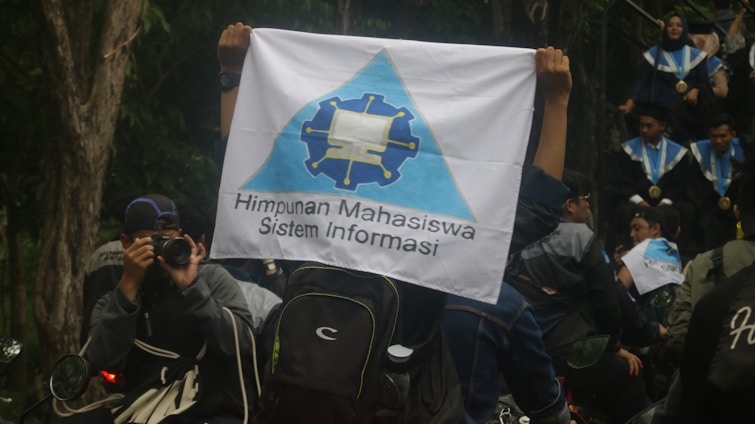 A person is holding a flag with a logo and the text 'Himpunan Mahasiswa Sistem Informasi'. People in graduation gowns and medals are gathered, with one individual photographing the scene. The environment is outdoors with trees visible in the background.