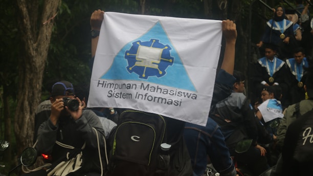 A person is holding a flag with a logo and the text 'Himpunan Mahasiswa Sistem Informasi'. People in graduation gowns and medals are gathered, with one individual photographing the scene. The environment is outdoors with trees visible in the background.