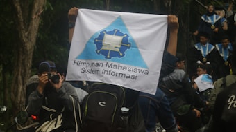A person is holding a flag with a logo and the text 'Himpunan Mahasiswa Sistem Informasi'. People in graduation gowns and medals are gathered, with one individual photographing the scene. The environment is outdoors with trees visible in the background.