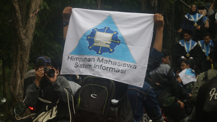 A person is holding a flag with a logo and the text 'Himpunan Mahasiswa Sistem Informasi'. People in graduation gowns and medals are gathered, with one individual photographing the scene. The environment is outdoors with trees visible in the background.