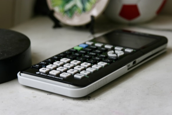 A scientific calculator with a black and white keyboard is placed on a desk. In the background, a round object resembling a soccer ball and a decorative green plate are visible.