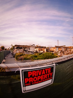 A chain-link fence with a 'Private Property No Trespassing' sign restricts access to an area with scattered debris and remnants of a demolished structure. The sky above is cloudy with a hint of blue.