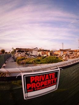 A chain-link fence with a 'Private Property No Trespassing' sign restricts access to an area with scattered debris and remnants of a demolished structure. The sky above is cloudy with a hint of blue.