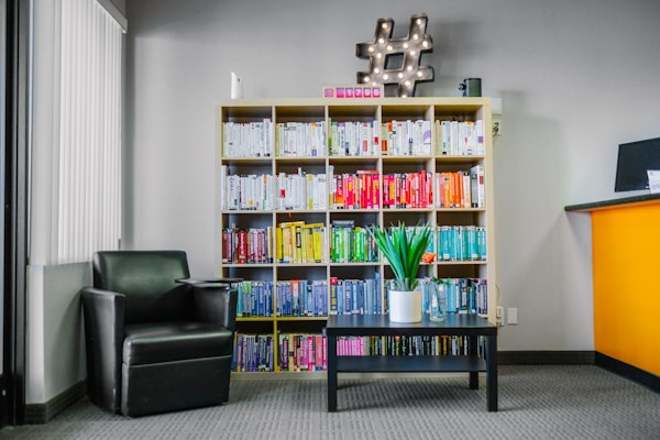 A modern interior space features a bookshelf filled with color-coded books, a black leather armchair next to a small black coffee table with a green potted plant. A decorative hashtag symbol with lights rests on top of the bookshelf. The walls are painted gray and a partial view of a bright orange reception desk is visible on the right.