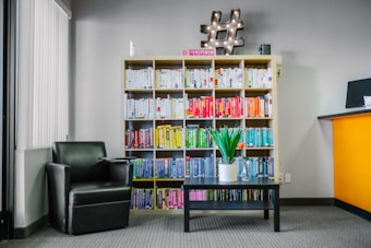 A modern interior space features a bookshelf filled with color-coded books, a black leather armchair next to a small black coffee table with a green potted plant. A decorative hashtag symbol with lights rests on top of the bookshelf. The walls are painted gray and a partial view of a bright orange reception desk is visible on the right.