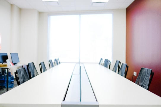 A spacious conference room with a long table and several chairs arranged neatly on both sides. The wall to the right is painted maroon, while the wall to the left is a light beige. Natural light enters the room through a large window at the far end. Several computer monitors are visible on the left corner of the room.