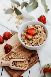 A white bowl filled with granola and sliced strawberries sits on a wooden board. Surrounding the bowl are whole strawberries, scattered granola, and a small wooden scoop filled with granola. Eucalyptus leaves and a white cloth add a natural touch to the scene.