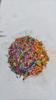 A pile of multicolored crayon shavings is scattered on a white surface. The shavings include vibrant hues of red, blue, yellow, green, pink, and orange, creating a visually striking mix of colors. The surrounding area shows slight smudges and marks from the shavings.