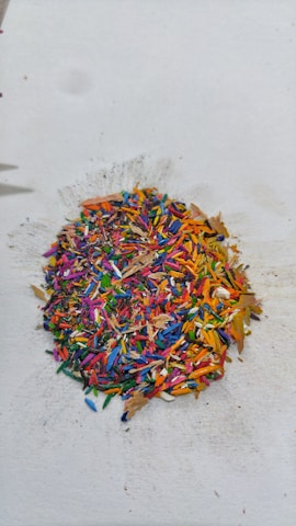 A pile of multicolored crayon shavings is scattered on a white surface. The shavings include vibrant hues of red, blue, yellow, green, pink, and orange, creating a visually striking mix of colors. The surrounding area shows slight smudges and marks from the shavings.