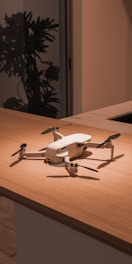 A white drone is placed on a wooden surface in an indoor setting. The background features a dark silhouette of a plant and a door or window frame, suggesting a modern interior.