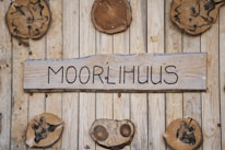 A wooden wall with several round, wooden slices attached, featuring unique dark patterns. In the center, there is a rectangular wooden plaque with the word 'MOORLIHUUS' written in a bold, black font.